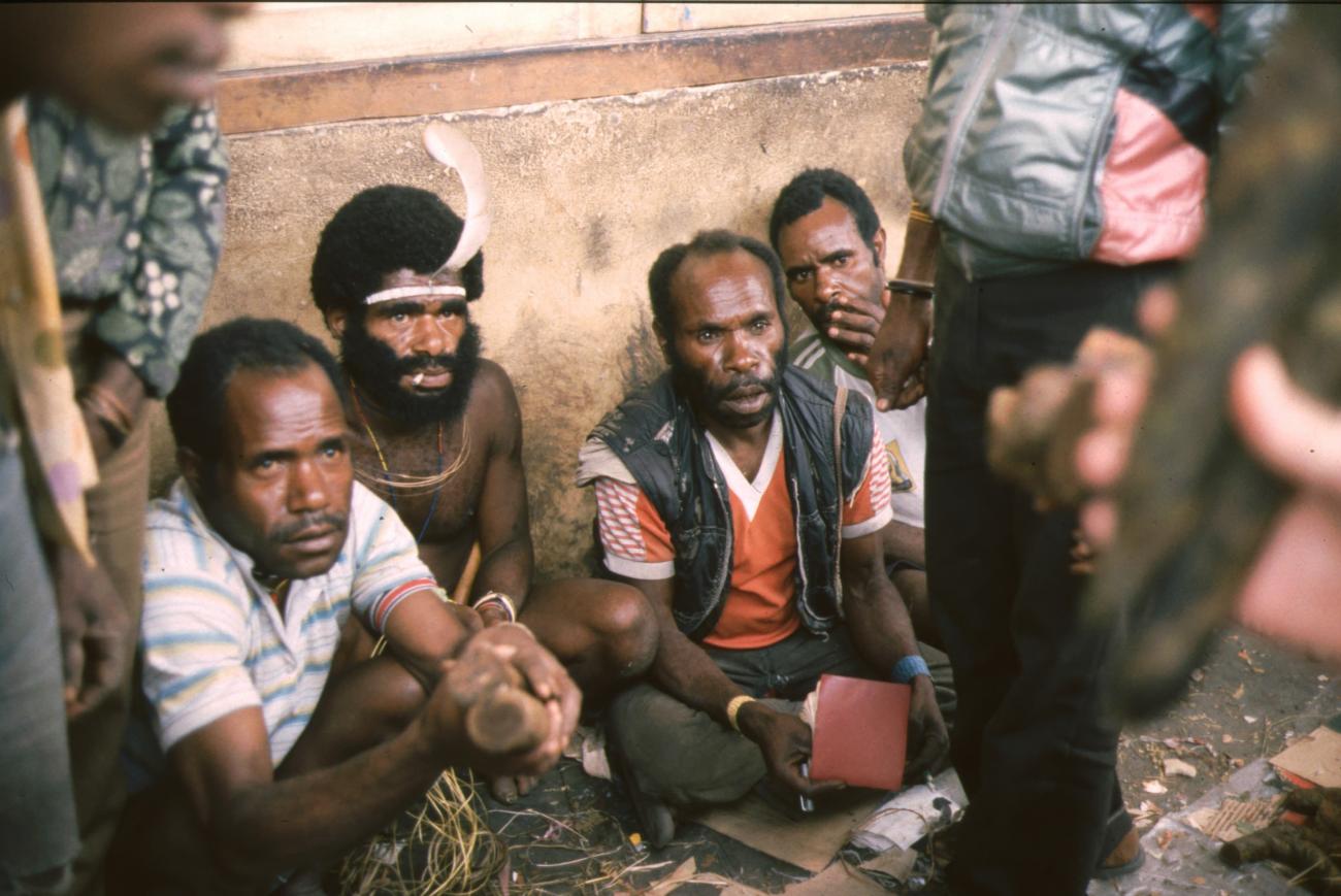 BD/166/282 - 
Papuamannen in westerse en traditionele kleding, zittend tegen een muur
