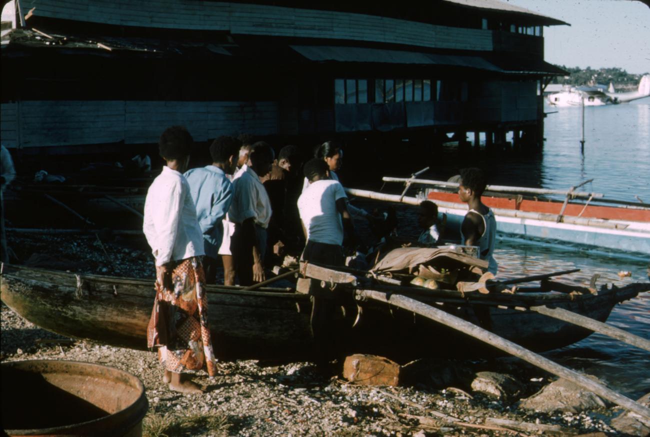 BD/288/95 - 
Groep papoea&#039;s bij prauw aan de haven met op de achtergrond een watervliegtuig

