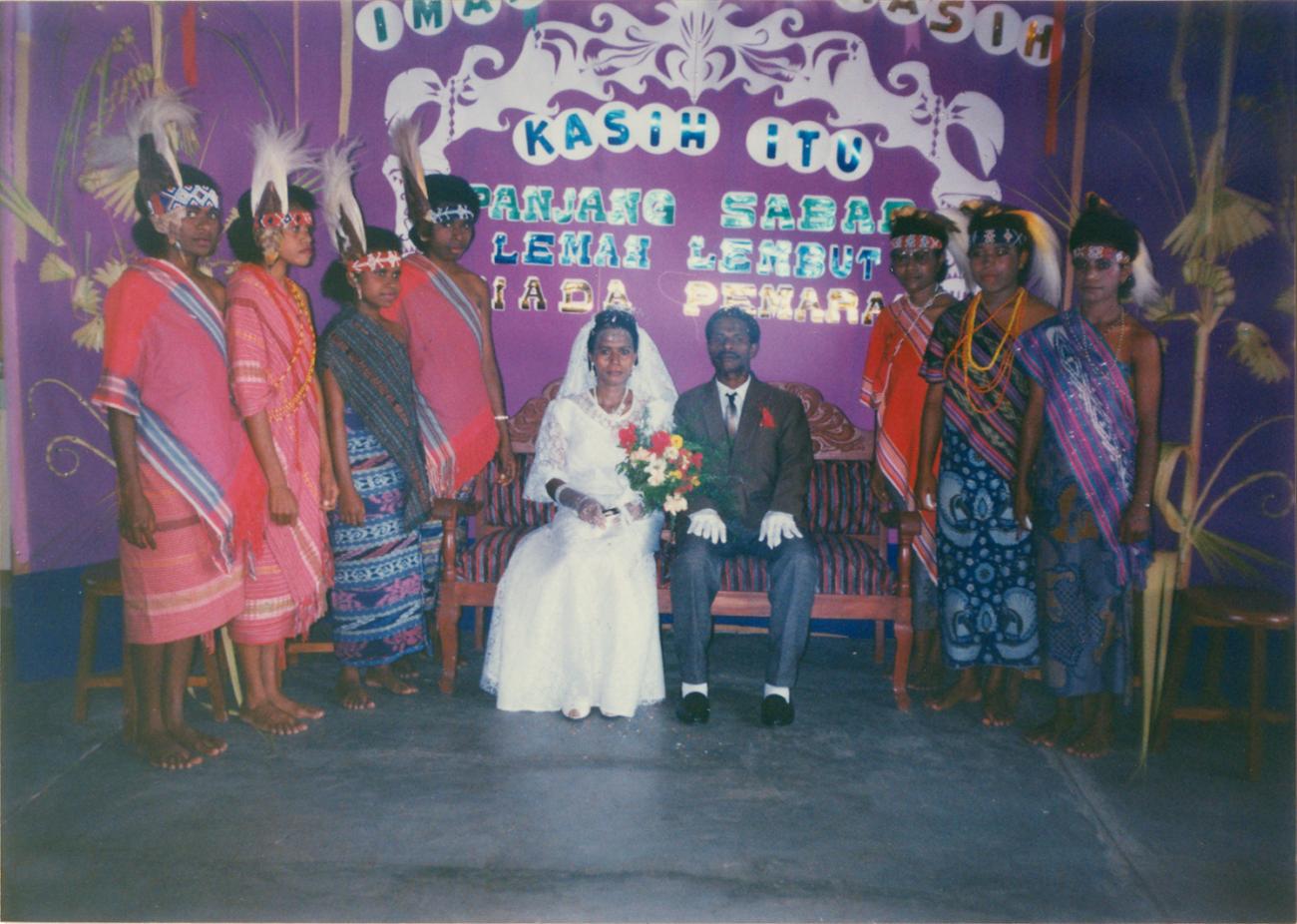 BD/309/53 - 
Dorp Teminabuan, bruid en bruidegom op bank met naast zich traditioneel geklede jonge vrouwen
