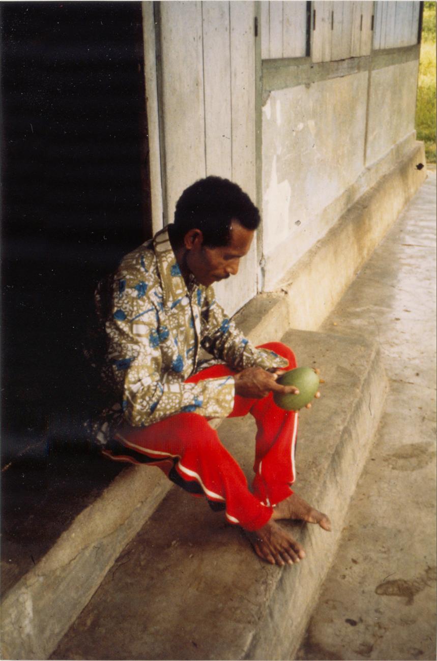 BD/309/66 - 
Dorp Teminabuan, man zittend bij ingang gebouw met vrucht in handen
