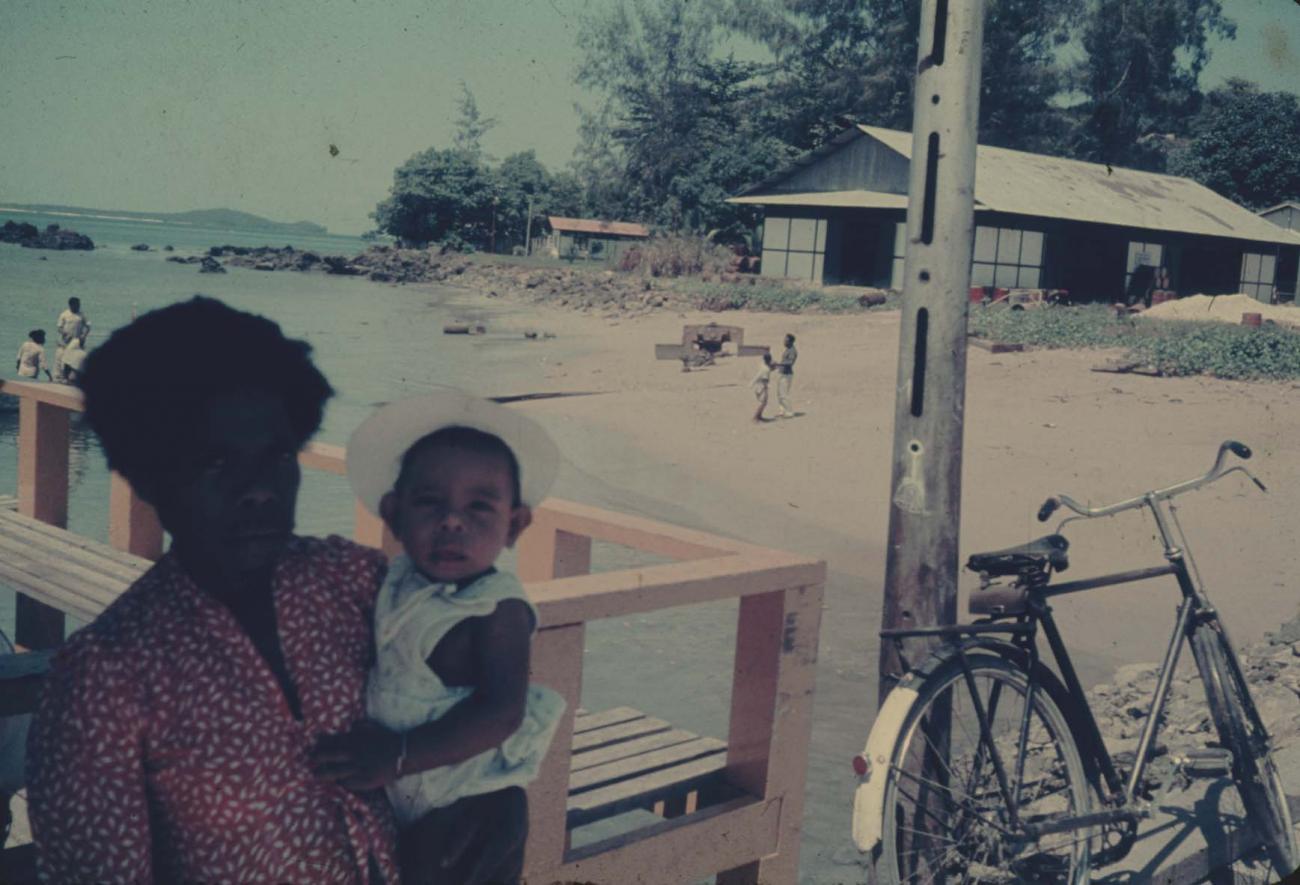 BD/144/257 - 
Vrouw met kind op arm, kust op achtergrond
