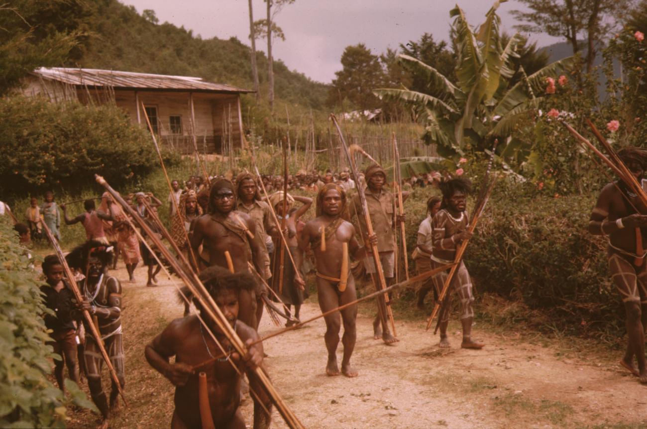 BD/132/153 - 
mannen in traditionele kleding met pijlen boog en speer gevolgd door dorpelingen
