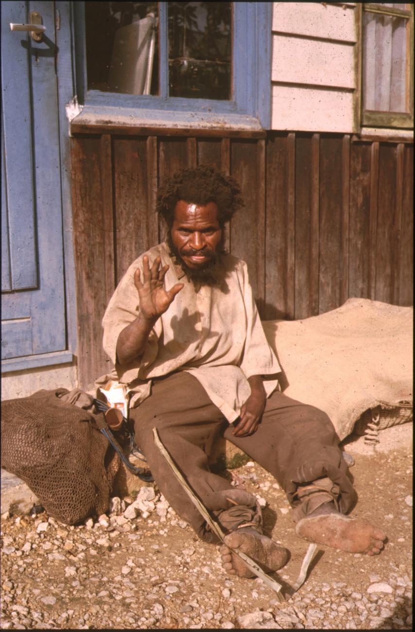 BD/132/24 - 
portretfoto van man zittend op de grond voor huis
