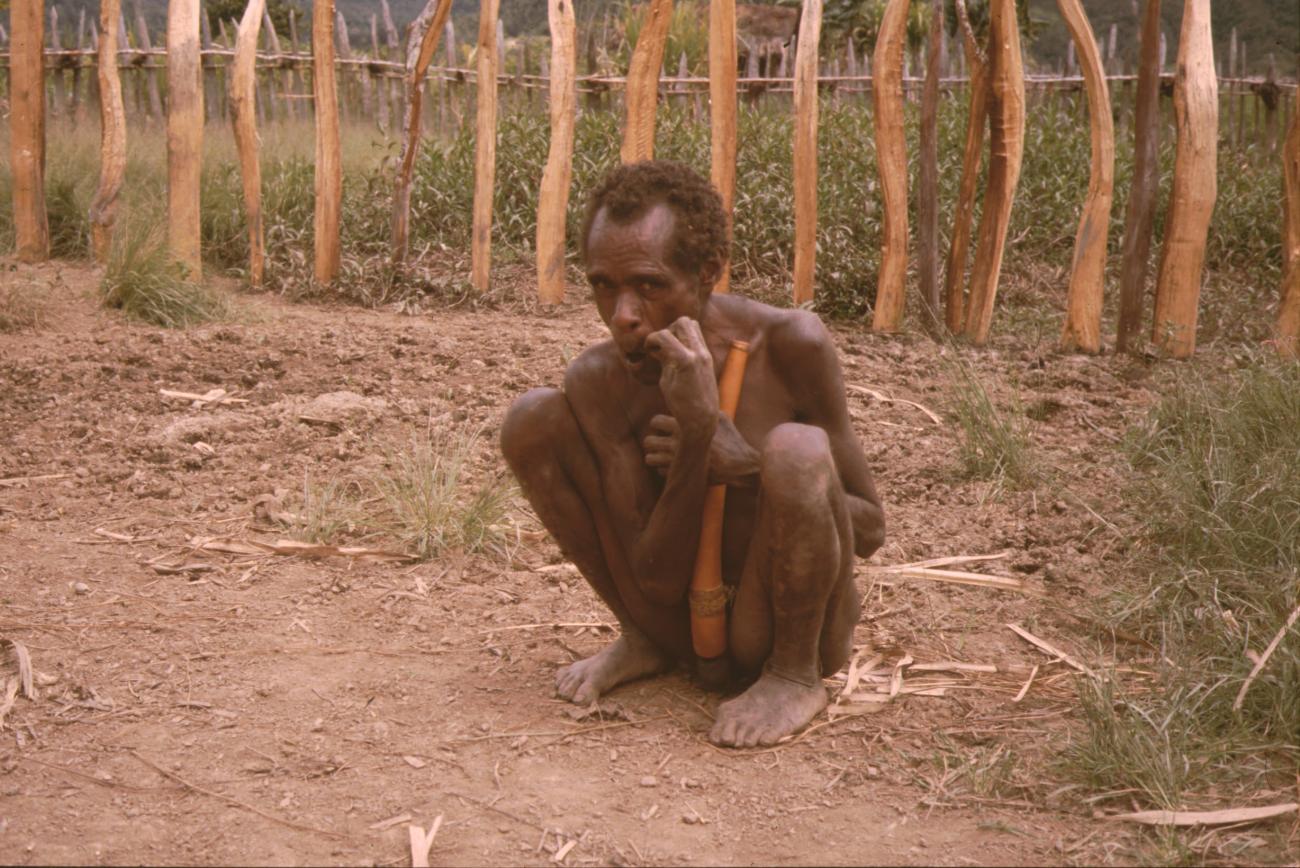 BD/132/73 - 
hurkende man met peniskoker voor omheinde tuin

