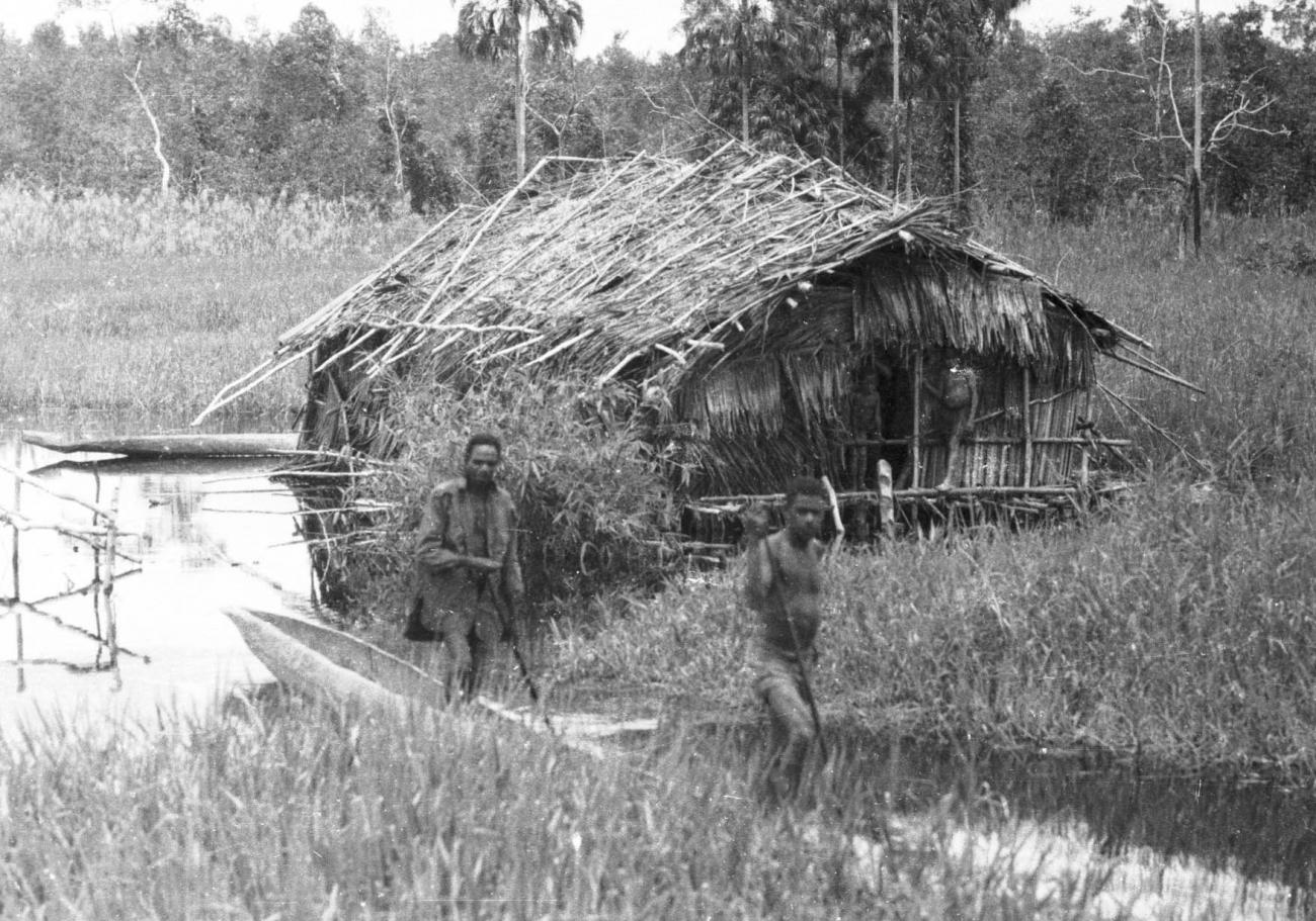 BD/133/421 - 
Tocht Merauke-Kepi-Cookrivier vv: Bemande prauw bij een hut aan de rivier
