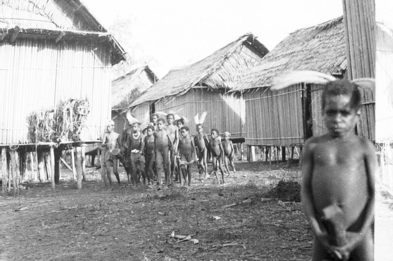BD/133/448 - 
Tocht Merauke-Kepi-Cookrivier vv: Portret van een jongen met paradijsvogelveren als hoofdsieraad en bamboekoker in zijn handen, met dorpsbewoners op achtergrond
