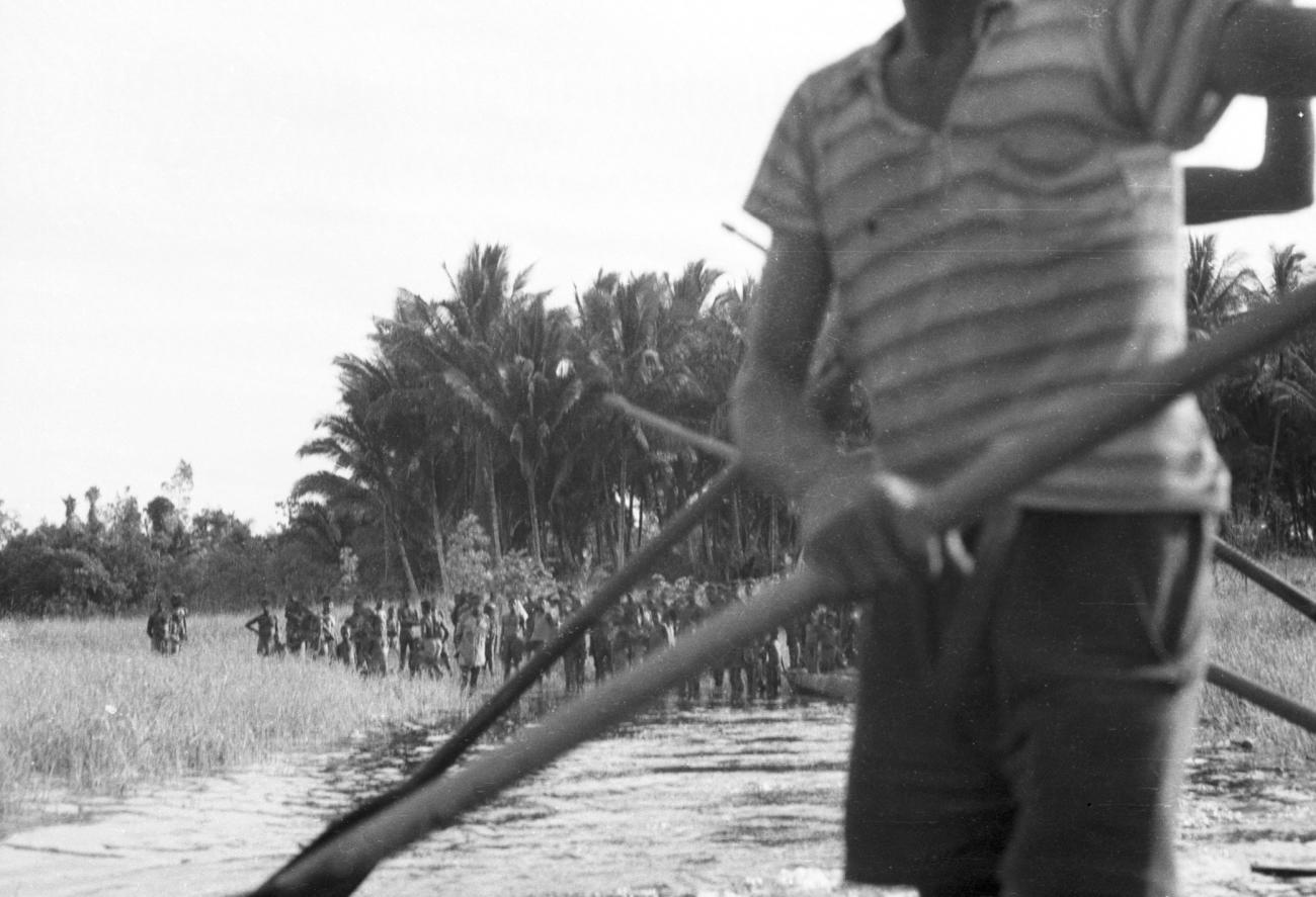 BD/133/657 - 
Zicht op rivieroever met dorpelingen en palmbomen vanaf prauw

