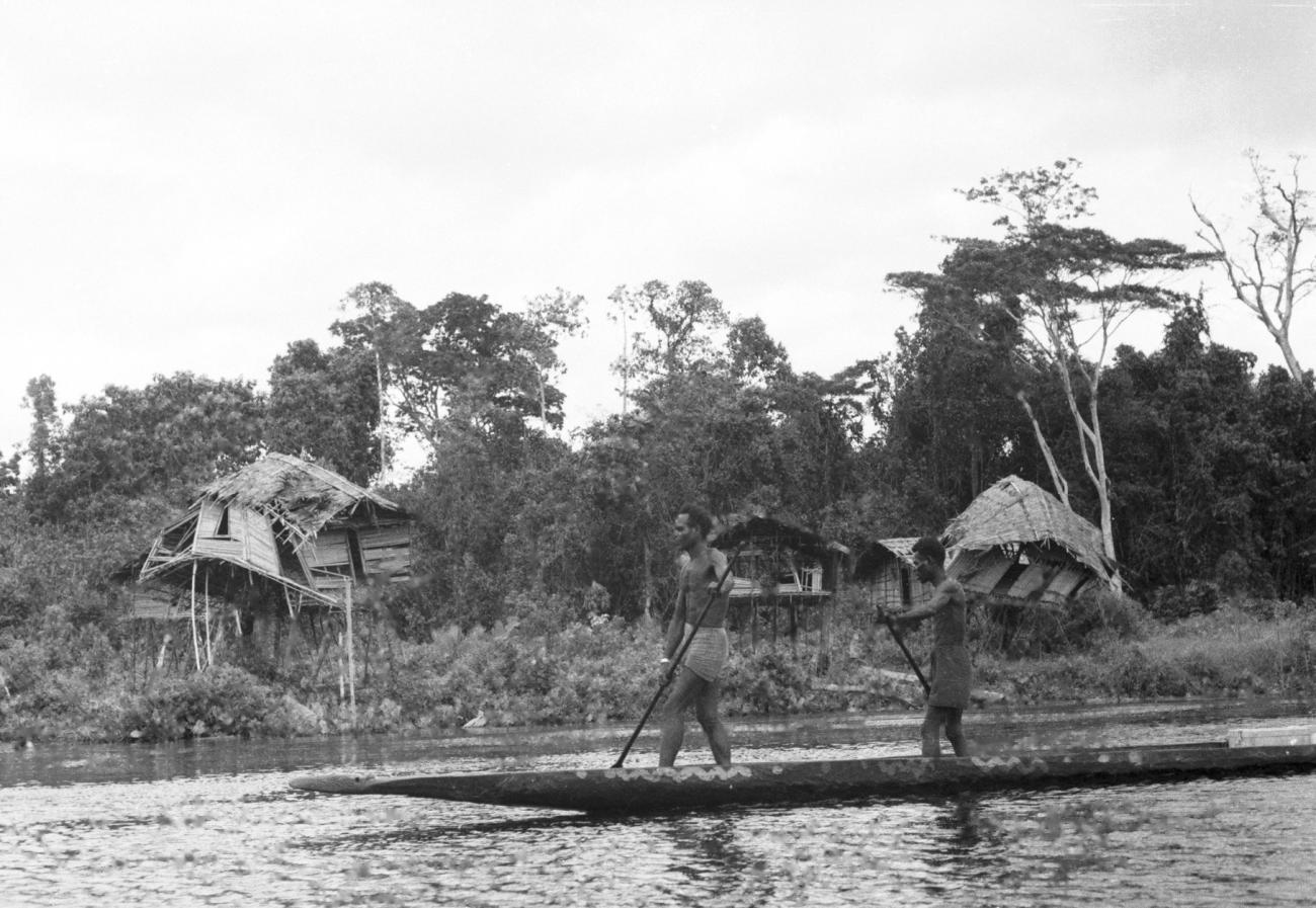 BD/133/665 - 
Prauw met Papoea-mannen, op de oever vervallen paalhutten
