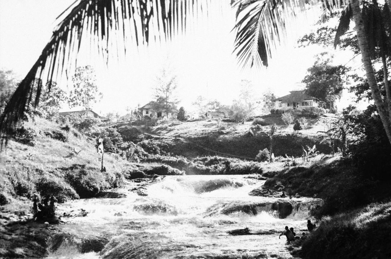 BD/133/679 - 
Stroomversnelling in de rivier met nederzetting op de hoge oever en Papoea&#039;s langs het water
