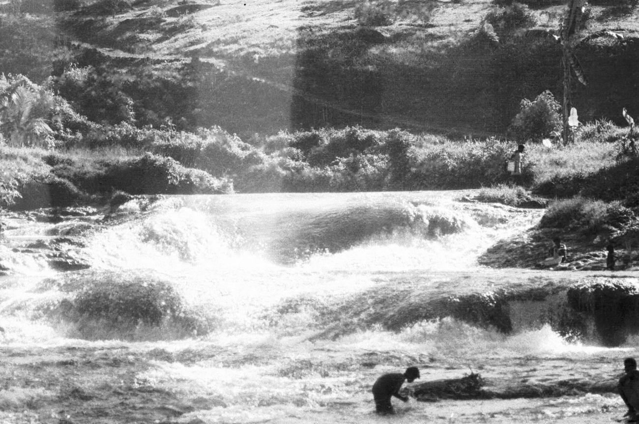 BD/133/680 - 
Stroomversnelling met hoge rivieroever en Papoea-vrouwen en kinderen bezig langs het water
