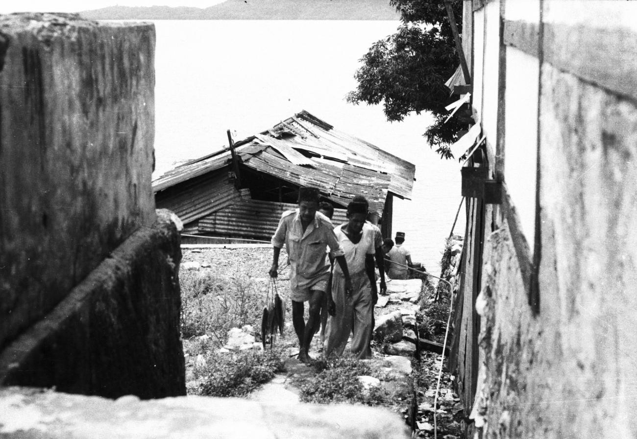 BD/133/910 - 
Een echtpaar beklimt de steile rivieroever bij een nederzetting, koppel vissen in de hand
