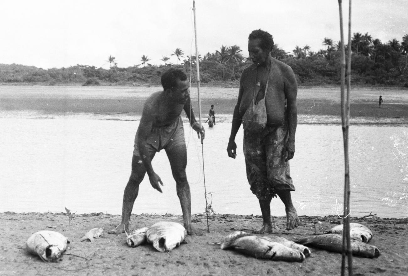BD/133/952 - 
Papoeaman en -vrouw met gevangen vis en vistuig aan de oever
