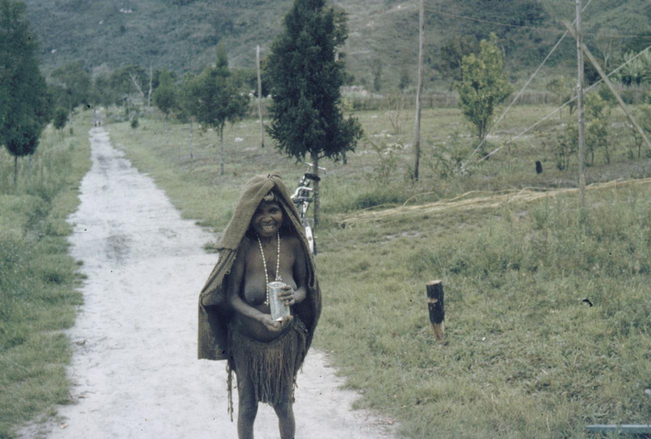 BD/285/84 - 
Vrouw met flesje in haar hand en doek om hoofd en ketting om hals poseert op landweg
