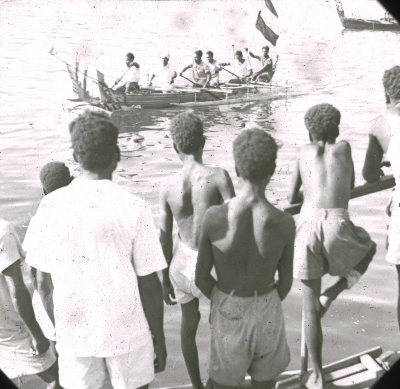 BD/329/13 - 
Groep Papoea-jongeren bekijkt vanaf de oever een prauw met dubbele uitleggers en versierde voorsteven
