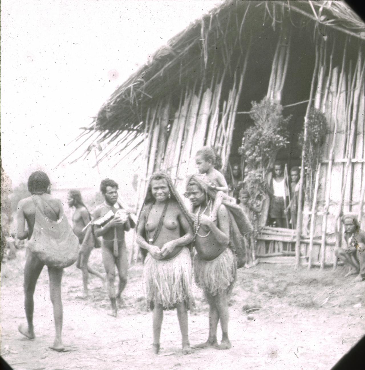 BD/329/18 - 
Papoea-mannen en -vrouwen met peniskokers en grasrokjes, voor een groot, met takken versierd huis
