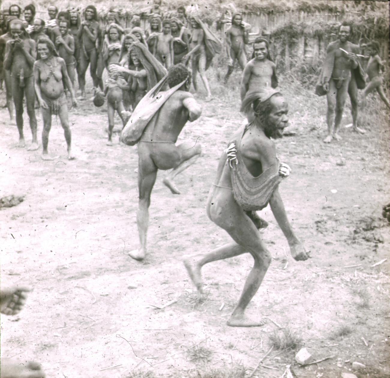 BD/329/27 - 
Twee Papoea-mannen voeren een dans uit onder toezien van stamgenoten
