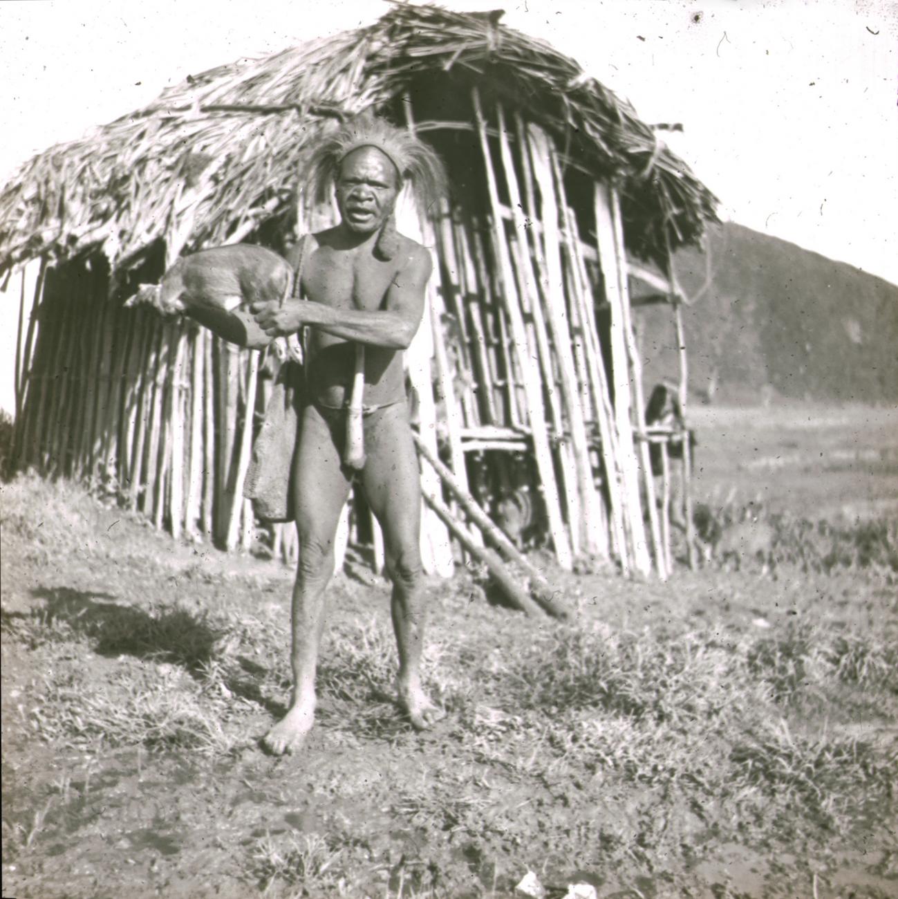 BD/329/44 - 
Papoea-man poseert voor een hut met een varkentje in zijn hand
