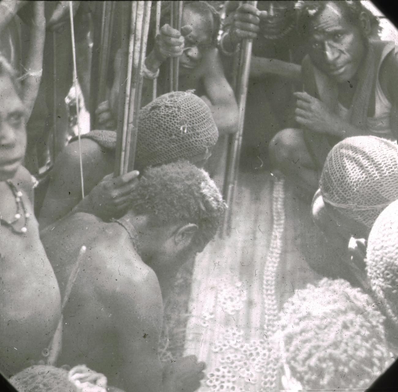 BD/329/4 - 
Papoea-mannen met hoofdversiering, draagnetten en pijl-en-boog, bezig met spel of handwerk 

