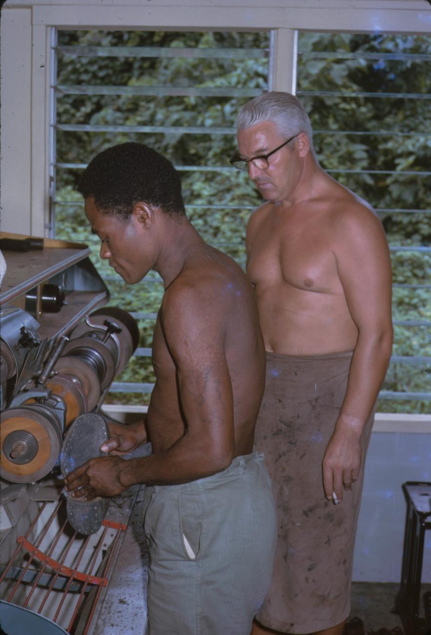 BD/209/9210 - 
Opleidingskamp Papoea Vrijwilligers Korps: leerling slijpt schoenzool bij schoenmakerijmachine 
