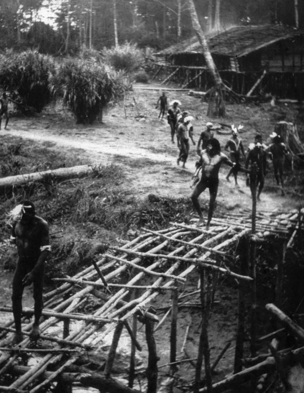 BD/216/118 - 
Mannen lopen op een oude voetbrug van stammetjes over een water
