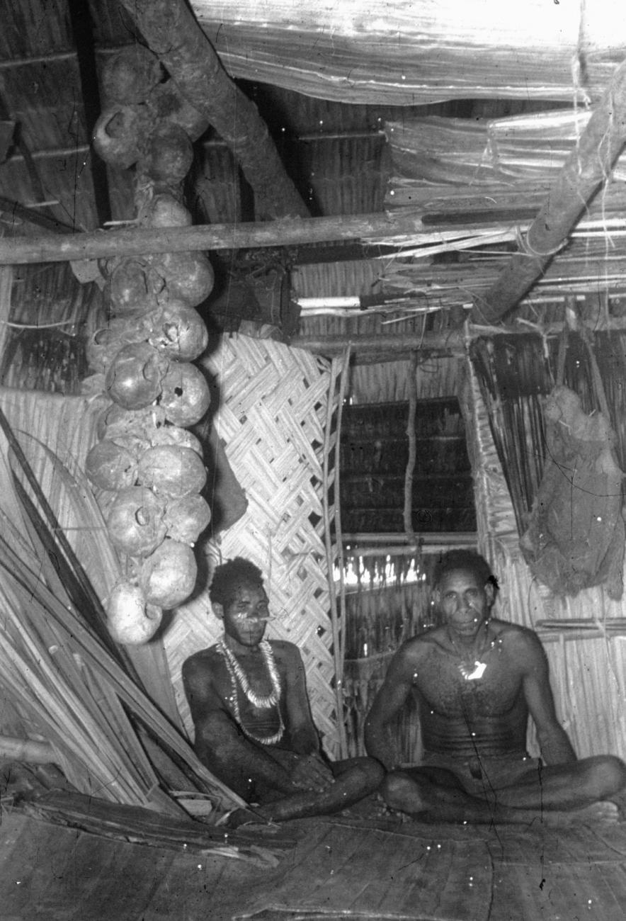 BD/216/205 - 
Twee Asmatters in het mannenhuis met een streng schedels hangend aan plafond
