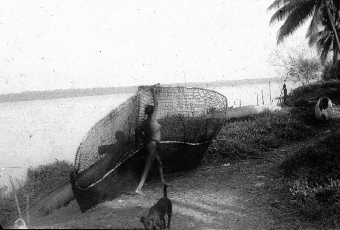 BD/216/245 - 
Vrouw draagt visnet langs de rivieroever 
