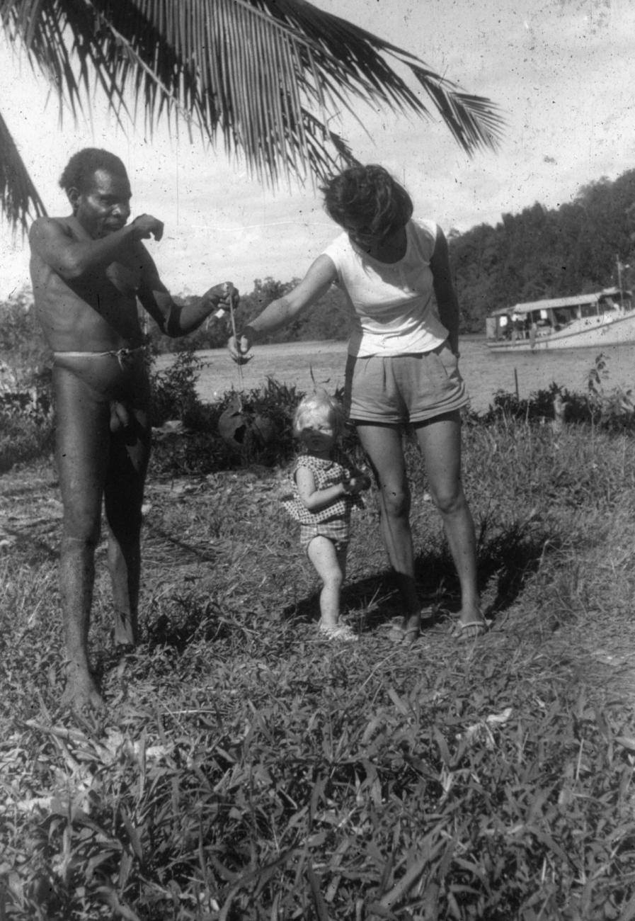 BD/216/287 - 
Man toont schedel aan blanke vrouw en kind aan oever van rivier met gouvernementsvaartuig
