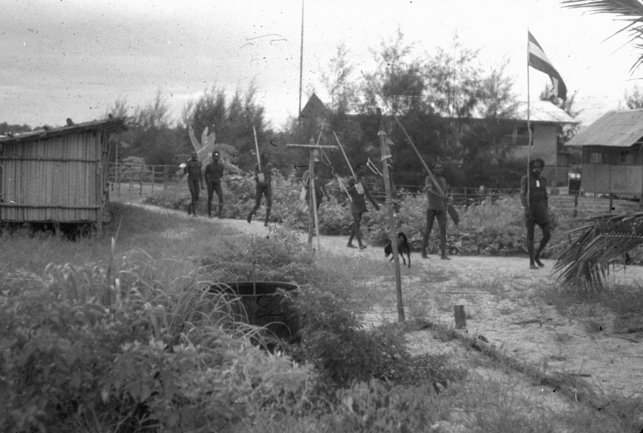 BD/216/307 - 
Asmatters lopen door het dorp met de Nederlandse vlag
