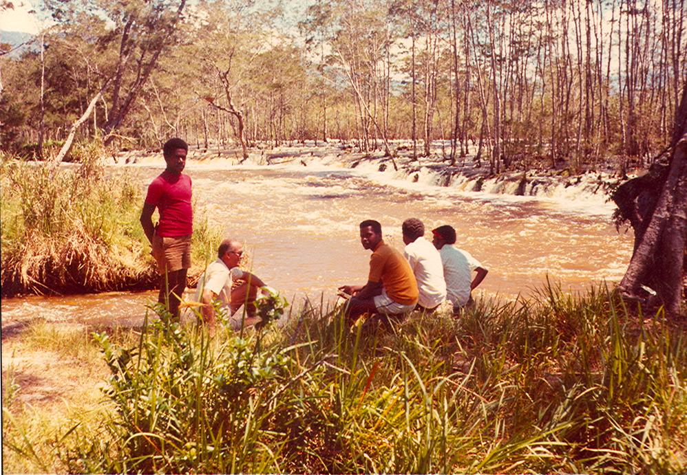 BD/269/144 - 
Broeder Henk Blom o.f.m. met Papoea&#039;s bij een buiten de oever getreden rivier
