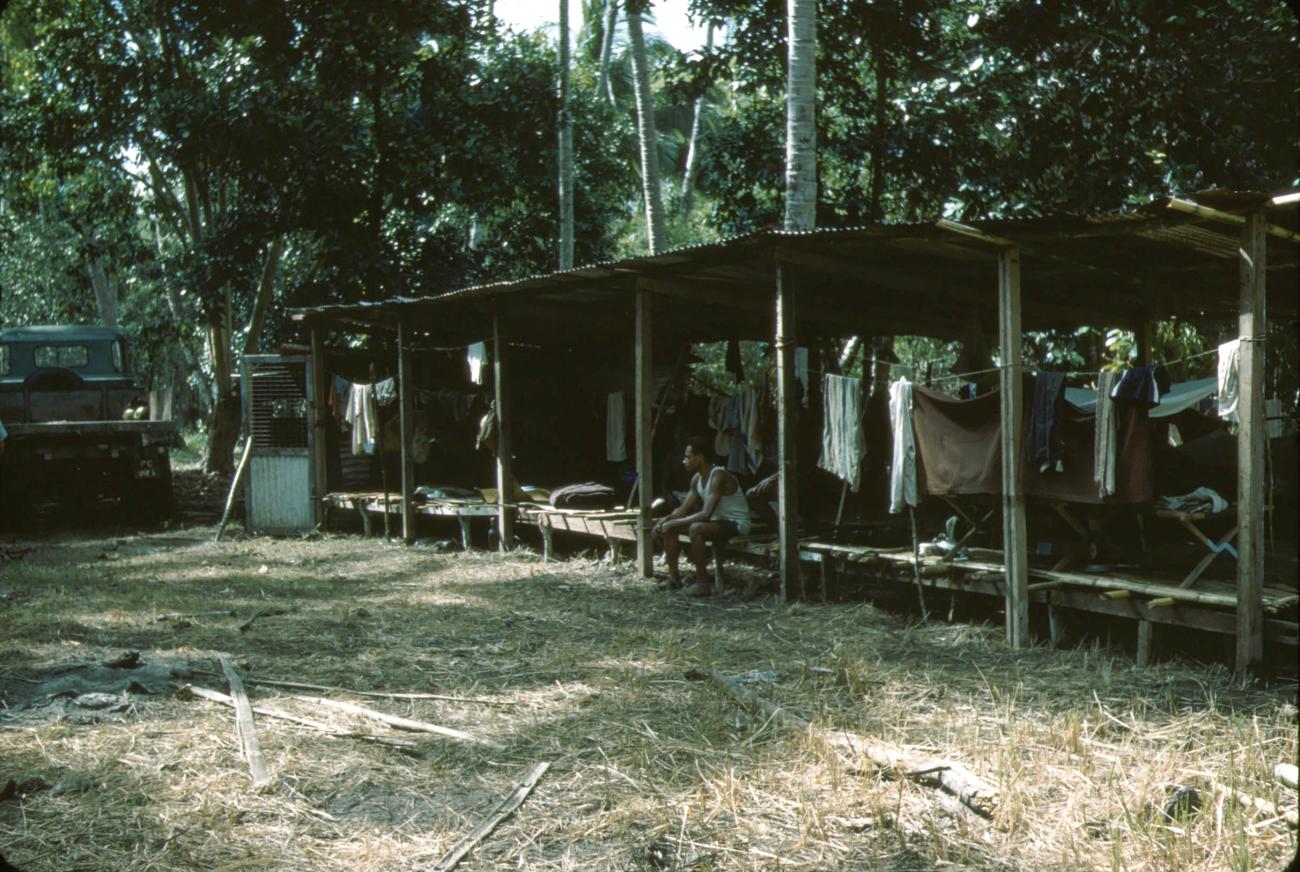 BD/288/168 - 
Man zittend in eenvoudig houten gebouw in bos
