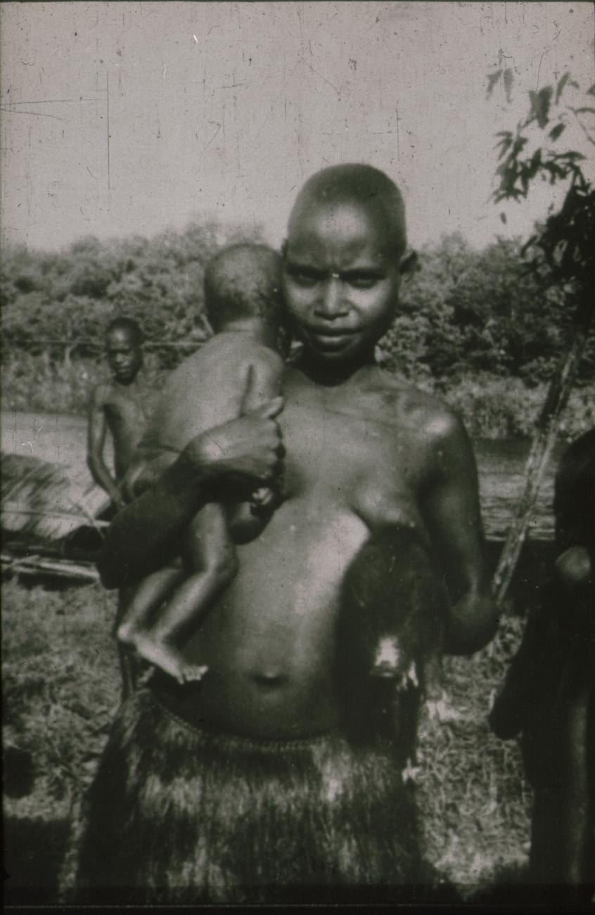 BD/309/215 - 
Vrouw in rieten rok met kind en varkentje, man en prauw op de achtergrond
