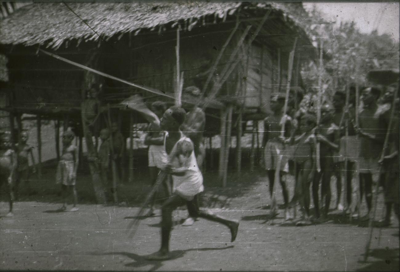 BD/309/254 - 
Mannen gooien met speer voor een traditionele paalwoning
