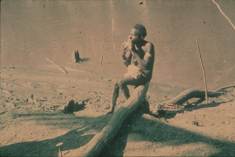 BD/30/50 - 
Man zit te eten op boomstam aan rivieroever
