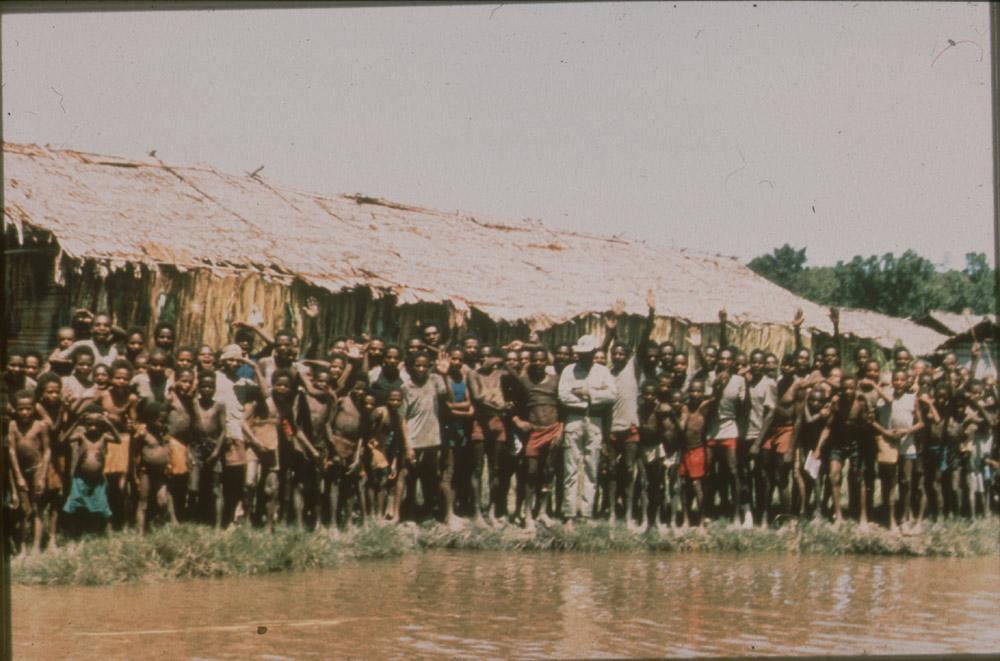 BD/30/90 - 
Asmatters staan te zwaaien op oever voor hun dorp
