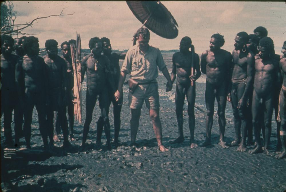 BD/30/99 - 
Bestuursambtenaar staat onder parasol met Asmatmannen op modderig strand
