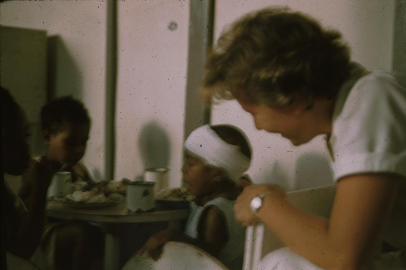 BD/171/512 - 
Verpleegster kijkt naar etende kimderen.
