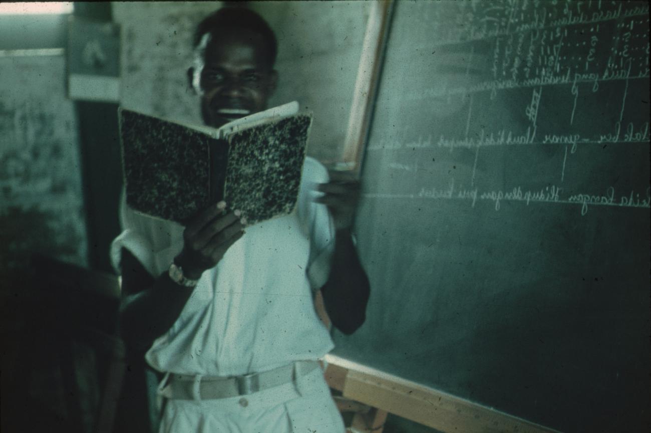 BD/171/582 - 
Man staat voor schoolbord met boek in handen.
