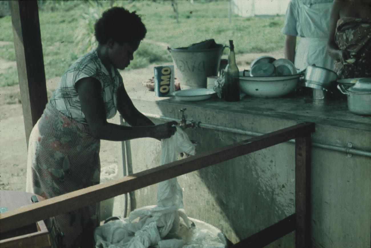 BD/171/583 - 
Vrouw wast kleren bij kraan.
