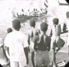 BD/329/13 Groep Papoea-jongeren bekijkt vanaf de oever een prauw met dubbele uitleggers en versierde voorsteven