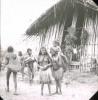 BD/329/18 Papoea-mannen en -vrouwen met peniskokers en grasrokjes, voor een groot, met takken versierd huis